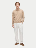 A man modeling a beige long-sleeve knit polo sweater, paired with white trousers and brown leather shoes, against a solid light gray background.