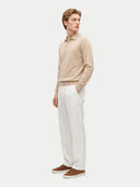 A man wearing a beige long-sleeve polo sweater, white trousers, and brown suede sneakers, standing in profile against a light gray studio background.