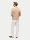 A man shown from the back wearing a light beige knit sweater, white trousers, and brown slip-on shoes against a plain light gray studio background.