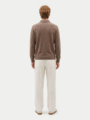 A man viewed from behind wearing a brown knit polo sweater and off-white trousers, standing against a light gray studio background.