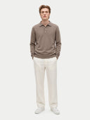 A male model stands against a light gray studio background wearing a taupe-brown long-sleeve polo sweater, off-white trousers, and white sneakers.