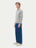 A man wearing a light gray long-sleeve polo sweater, wide-leg blue jeans, and off-white sneakers, standing in profile against a plain light gray studio background.