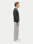 A man in profile wearing a dark charcoal gray long-sleeve crewneck sweater, light gray wide-leg sweatpants, and white sneakers, standing against a solid off-white background.