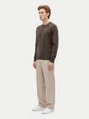 A man wearing a dark brown crewneck sweater, beige wide-leg trousers, and white sneakers, standing in profile against a light gray studio background.