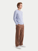 A man wearing a light blue cable-knit sweater, brown trousers, and white sneakers, standing in profile against a light gray studio background.