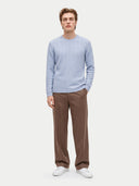 A man modeling a light blue cable-knit sweater and brown trousers against a plain light gray studio background.