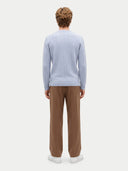 A man viewed from behind wearing a light blue cable-knit sweater, brown trousers, and white sneakers against a plain light gray studio background.