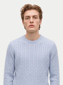 A young man with light brown hair wearing a light blue cable-knit crewneck sweater, photographed against a solid off-white background.