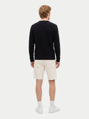 A man viewed from the back wearing a black cable-knit sweater, off-white shorts, white socks, and white sneakers against a light gray studio background.