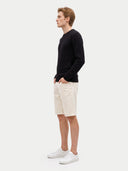 A man in profile wearing a black cable-knit sweater, off-white shorts, and white sneakers, standing against a light gray studio background.