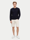 A man modeling a dark navy blue cable-knit sweater, cream-colored shorts, and white sneakers against a light gray studio background.