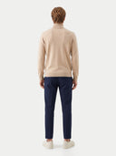 A man viewed from the back wearing a beige mock-neck knit sweater, navy blue trousers, and off-white sneakers, set against a plain light gray studio background.