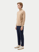 A man wearing a beige turtleneck sweater, navy blue trousers, and off-white sneakers, standing in profile against a light gray studio background.