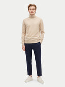 A man modeling a beige ribbed turtleneck sweater, navy blue trousers, and off-white sneakers against a light gray studio background.