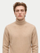 A man wearing a light beige knit turtleneck sweater, photographed against a plain light gray studio background.