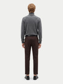 A man viewed from the back wearing a gray turtleneck sweater, dark brown trousers, a black belt, and black shoes, standing against a light gray studio background.