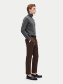 A man in a side profile wearing a dark gray turtleneck sweater, brown trousers, and black leather loafers, standing against a light gray studio background.