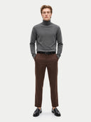 A man modeling a dark gray turtleneck sweater, brown trousers, a black belt, and black leather loafers, standing against a light gray studio background.