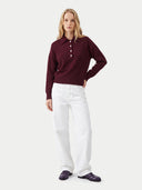 A woman modeling a burgundy long-sleeve polo knit top with white buttons, paired with white trousers and dark purple loafers, against a light gray studio background.
