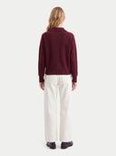 A woman viewed from the back wearing a burgundy knit mock-neck sweater and off-white wide-leg trousers, standing against a light gray studio background.