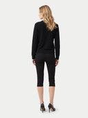 A woman viewed from the back wearing a black long-sleeve top, black capri pants, and black heels against a light gray studio background.