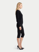 A woman in a black long-sleeve top and black capri pants stands in profile against a light gray studio background, wearing black pointed-toe heels.