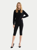 A woman modeling a black long-sleeve crewneck sweater and matching black capri leggings with black high heels, set against a plain light gray studio background.