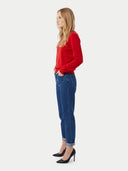 A woman in a red long-sleeve top, blue jeans, and black heels, shown in profile against a light gray studio background.