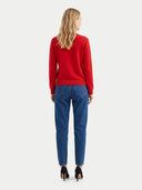 A woman viewed from the back wearing a bright red long-sleeve sweater, blue jeans, and black high heels against a light gray studio background.