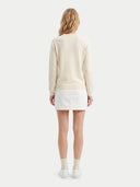A woman viewed from behind wearing a cream-colored long-sleeve knit sweater, a white mini skirt, white socks, and white sneakers against a light gray studio background.