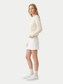 A woman modeling a cream-colored long-sleeve knit sweater, white mini skirt, white socks, and white sneakers against a light gray studio background.