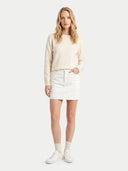 A woman modeling a cream-colored long-sleeve knit sweater and a white mini skirt, paired with white socks and white sneakers, against a solid light gray background.