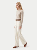 A woman modeling a light beige cable-knit sweater, off-white wide-leg trousers, a brown belt, and brown leather shoes against a plain light gray studio background.