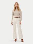 A woman stands against a light gray studio background wearing a beige cable-knit sweater, off-white wide-leg trousers, a brown belt, and brown ankle boots.