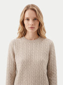 A woman with blonde hair wearing a beige cable-knit crewneck sweater, photographed against a clean light gray studio background.