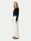 A woman stands in profile wearing a black ribbed long-sleeve top, off-white wide-leg trousers, a brown belt, and brown shoes against a light gray studio background.