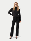 A woman modeling a black long-sleeve turtleneck sweater and matching black flared trousers with black pointed-toe heels, photographed against a light gray studio background.