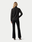 A woman shown from the back wearing a dark charcoal gray mock neck sweater and black flared trousers, standing against a light gray studio background.