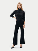 A woman modeling a black long-sleeve turtleneck sweater and matching black wide-leg trousers, styled with black pointed-toe heels against a light gray studio background.