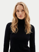 A blonde woman wearing a black ribbed turtleneck sweater, photographed against a clean light gray studio background.