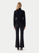 A woman viewed from the back wearing a black long-sleeve mock-neck top and matching black flared pants, standing against a light gray studio background.