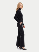 A woman modeling a black long-sleeve mock-neck top and matching black flared pants with black pointed-toe heels, photographed against a plain light gray studio background.