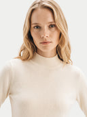 A blonde woman wearing a cream-colored ribbed mock neck sweater, photographed against a clean, light off-white background.