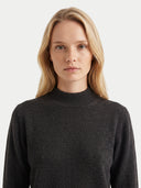 A blonde woman wearing a dark charcoal gray mock neck knit sweater, photographed against a clean light gray background.