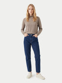 A woman modeling a beige mock-neck knit sweater and dark blue straight-leg jeans with off-white sneakers, standing against a light gray studio background.