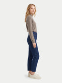 A woman stands in profile wearing a light taupe knit mock-neck sweater, dark blue cropped jeans, and off-white sneakers against a plain light gray studio background.