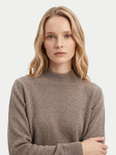 A blonde woman wearing a light brown ribbed mock neck sweater, photographed against a clean, light gray studio background.