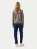 A woman viewed from behind wearing a taupe knit sweater, dark blue jeans, and off-white sneakers against a light gray studio background.