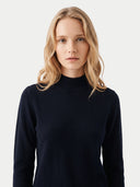 A blonde woman wearing a dark navy blue mock neck sweater, photographed against a clean, light gray studio background.