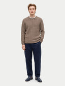 A man modeling a taupe cable-knit crewneck sweater, paired with navy blue trousers and dark loafers, against a light gray studio background.
